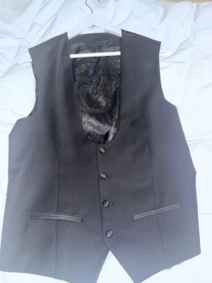 Svart kostymväst med paisleymönster - Snygg svart kostymväst med fem knappar framtill och två fickor. Baksidan har ett glansigt paisleymönstrat tyg och justerbart spänne. Perfekt att kombinera med skjorta för en stilren look. Materialet känns lyxigt och har en elegant finish.