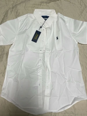 Vit kortärmad skjorta Ralph Lauren - Klassisk vit kortärmad skjorta från Ralph Lauren i 100% bomull. Skjortan har button-down krage, broderad marinblå logga på bröstet och knappar framtill. Passformen är classic fit och materialet känns svalt och fräscht. Perfekt till jeans eller chinos.