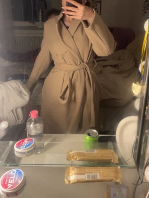 Beige kappa från Boohoo med bälte - Säljer en snygg beige kappa från Boohoo i storlek 34. Den har omlottmodell med bälte i midjan och långa ärmar. Perfekt för dig som gillar stilrena och klassiska plagg. Materialet känns mjukt och har en ull-liknande yta.