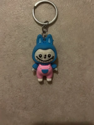 Ingen väst på nyckelring - Bilderna visar en nyckelring med en figur i blå huva och rosa byxor, men ingen väst syns på figuren. Ingen relevant information om väst kan ges.