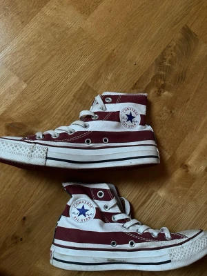 Converse USA-färger (höga) - Säljer ett par höga Converse med USA-flaggans design. Skorna har röda och vita ränder på sidorna och blå tunga med vita stjärnor. Klassisk rund tå, vit platt sula och snörning.