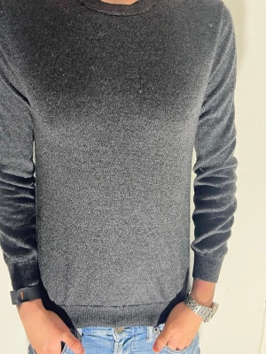 Mörkgrå merinotröja från Without Prejudice - Snygg mörkgrå tröja från Without Prejudice i 100% merinoull. Klassisk rund hals och långärmad modell med ribbade muddar. Perfekt för dig som gillar stilrena och mjuka plagg med skön passform. London-designad och tillverkad i Bangladesh.