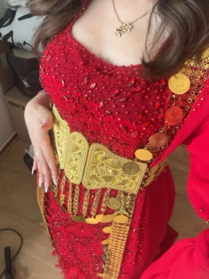 Kurdisk lawandi med guldiga detaljer - Säljer en röd långklänning dekorerad med glittriga paljetter och pärlor. Klänningen har en unik guldfärgad bälte och myntdetaljer längs sidorna. Lång ärm och fjäderliknande detaljer vid fållen ger extra elegans. Perfekt för speciella tillfällen. Passar 32-38. Pris går att diskuteras vid snabb affär.