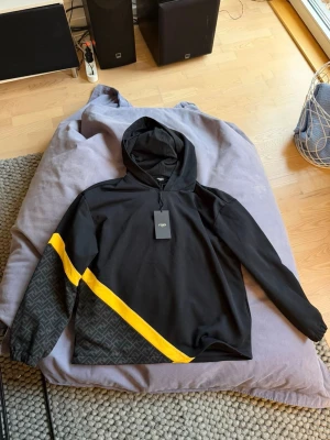 Svart Fendi hoodie med gul detalj - Aldrig använd! Snygg svart hoodie från Fendi med huva och en diagonal gul rand över bröstet och ärmen. Ena ärmen har Fendis klassiska monogrammönster i grått. Hoodien har en modern och sportig look, perfekt för dig som gillar streetwear och exklusiva märken. Nypris: 1300 euro