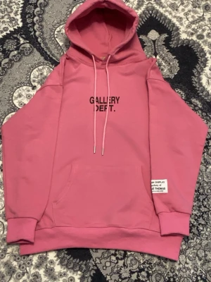 Rosa hoodie från Gallery Dept. - Säljer en rosa hoodie från Gallery Dept. med svart text på bröstet och en vit patch vid nederkanten. Hoodien har huva med dragsko, magficka och ribbade muddar. Perfekt för dig som gillar streetwear och vill sticka ut med färg.pris kan diskuteras.