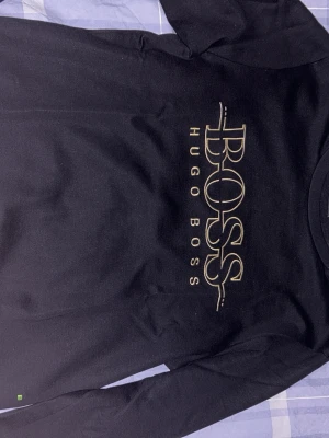 Svart långärmad tröja Hugo Boss - Svart långärmad tröja från Hugo Boss med stort guldigt logotryck på bröstet. Tröjan har rund halsringning och är tillverkad i mjukt material, perfekt för en stilren och avslappnad look.