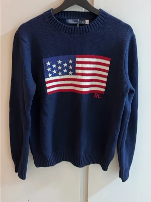 Mörkblå stickad tröja Polo Ralph Lauren - Mörkblå stickad tröja från Polo Ralph Lauren med amerikansk flagga i rött, vitt och blått på bröstet samt RL-broderi. Tröjan har rund halsringning och ribbade muddar. Perfekt för dig som gillar klassisk och sportig stil.