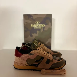 Valentino Rockrunner sneakers camouflage - Säljer ett par Valentino Rockrunner sneakers med snyggt kamouflagemönster i grönt, beige och brunt. Passar från 39-41  Skorna har detaljer i mocka och skinn, samt en röd häl med nitar. Platt sula och rund tå, med snörning framtill. Skicket är som på bild är i perfekt skick endast lite färg som försvunnit från framsidan av skon men knappts märks| medföljs dustbag box+ extra snören| fraktas den 2 januari 