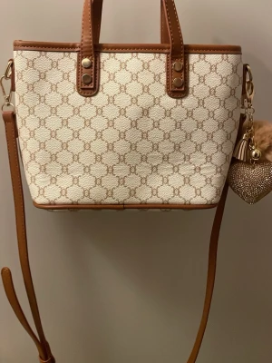 Elegant Vit & Beige handväska med monogrammönster - Snygg beige handväska med bruna detaljer och axelrem. Väskan har ett elegant monogrammönster i brunt och guldfärgade metalldetaljer. Klassisk fyrkantig form och dekorativt hjärthänge. Perfekt accessoar för dig som gillar stilrena väskor.