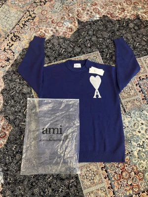  Ami Paris tröja - Säljer en blå stickad tröja från Ami Paris med deras ikoniska vita hjärta och bokstaven A på bröstet. Tröjan har rund halsringning, långa ärmar och ribbade muddar. Perfekt för dig som gillar stilrena och trendiga plagg.