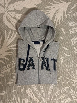 Grå zip-hoodie från GANT - Snygg grå hoodie från GANT med dragkedja och stor logga framtill. Modellen har huva med mörkblå snören och ribbade muddar. Tillverkad i mjuk bomullsblandning som känns skön mot huden. Perfekt för en avslappnad och sportig stil.