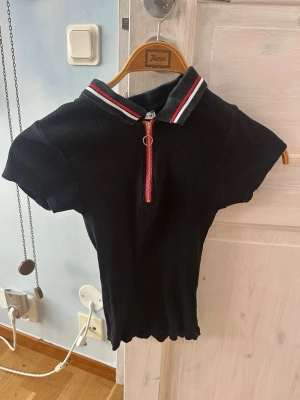 Svart ribbad polotopp från FB Sister - Svart ribbad polotopp från FB Sister med korta ärmar och dragkedja framtill. Kragen har röd, vit och grå randig detalj som ger en sportig vibe. Perfekt till jeans eller kjol för en trendig look.