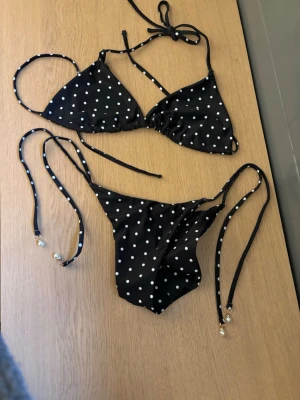Svart prickig bikini med pärldetaljer - Snygg svart bikini med vita prickar och tunna knytband. Både överdel och underdel har dekorativa pärlor på snörena. Klassisk trekantsmodell som ger en trendig och ungdomlig look, perfekt för stranden eller poolen. Aldrig använd!