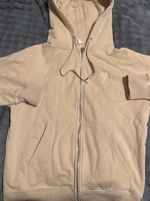 Beige hoodie med dragkedja från AMI - Snygg beige hoodie från AMI med dragkedja framtill och broderad logga på bröstet. Hoodien har huva med dragsko, ribbade muddar och två stora fickor fram. Tillverkad i mjuk bomullsblandning som känns riktigt skön mot huden.