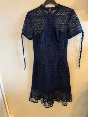 Marinblå spetsklänning med snörning - Snygg marinblå kortklänning i spets med korta ärmar och dekorativ snörning på ärmarna. Klänningen har rund halsringning och vackra spetsdetaljer över hela plagget. Perfekt för dig som vill ha en feminin och trendig look.