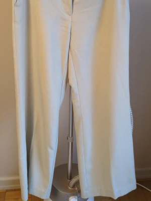 Vida beige kostymbyxor - Snygga beige kostymbyxor med hög midja och vida ben. Byxorna har bälteshällor och sidofickor, vilket ger en clean och modern look. Perfekta för dig som gillar minimalistisk stil och vill ha ett par byxor som sticker ut utan att vara för mycket.