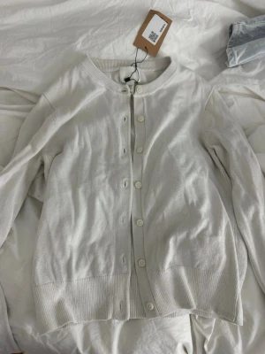 Vit kofta från H&M, XS - Snygg vit kofta från H&M i storlek XS. Koftan har rund halsringning, långa ärmar och knäppning framtill med vita knappar. Tillverkad i mjukt och tunt stickat material, perfekt för lager-på-lager-stil.