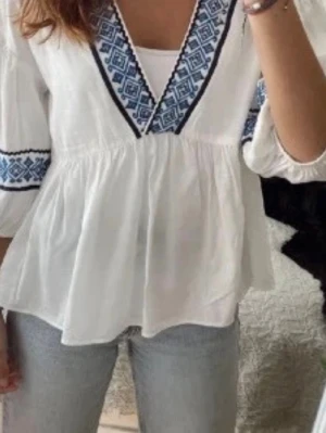 Vit blus med blått mönster - Snygg vit blus med blått etno-inspirerat mönster längs v-ringningen och ärmsluten. Blusen har en lös passform och trekvartsärmar. Perfekt att styla med jeans för en avslappnad look.