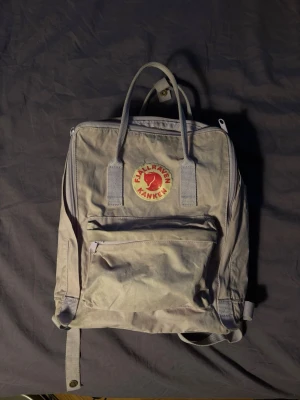 Ljusgrå Fjällräven Kånken ryggsäck - ny pris: 1299.                               Klassisk ljusgrå Fjällräven Kånken ryggsäck med det ikoniska runda röda och gula märket framtill. Ryggsäcken har ett stort huvudfack, en framficka med dragkedja och justerbara axelband. Tillverkad i slitstarkt syntetmaterial och har dubbla handtag upptill.