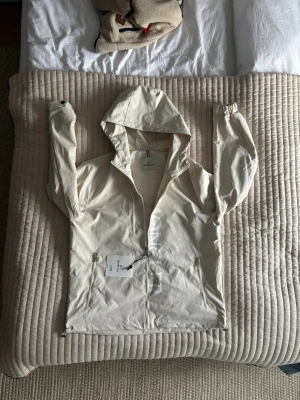 Vit vindjacka från Moncler - Stilren vit vindjacka från Moncler med huva och dragkedja framtill. Jackan har två sidofickor med dragkedja och diskret Moncler-logga längs dragkedjan. Lätt material som passar perfekt för blåsiga dagar. Storlek Xl men passar också som large