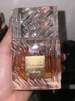 Khamrah parfym från Lattafa - Khamrah parfym från Lattafa i en elegant, fyrkantig glasflaska med mönstrad design och guldetikett. Flaskan har en transparent look med bärnstensfärgad vätska inuti och ett matchande mönstrat lock. Perfekt för dig som gillar lyxiga och unika dofter.
