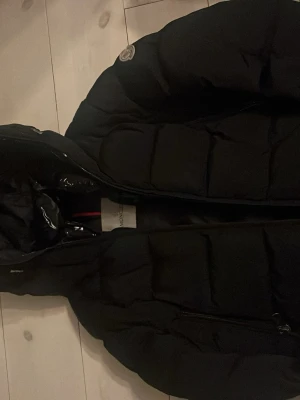 Svart dunjacka från Moncler - Säljer en svart dunjacka från Moncler med klassisk quiltad design och huva. Jackan har dragkedja framtill och en ikonisk Moncler-logga på axeln. Insidan har ett unikt serietidningsmönster som ger extra karaktär. Perfekt för kalla vinterdagar.
