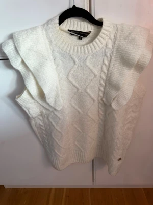 Vit stickad väst med volang - Vit stickad väst från Pernilla Wahlgrens kollektion. Snyggt kabelstickat mönster och breda volangdetaljer på axlarna. Rund halsringning och ribbade kanter. Perfekt för lager-på-lager och ger en trendig look.