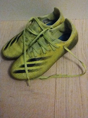Gula Adidas fotbollsskor med dobbar - Säljer ett par gula fotbollsskor från Adidas med svarta detaljer och klassiska ränder på sidan. Skorna har snörning och en ovandel i syntetmaterial med struktur för bättre bollkontroll. Yttersulan har fasta dobbar för spel på gräs. Stretchigt material som formar sig runt foten
