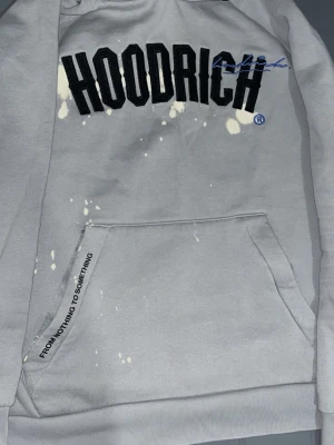Grå hoodie från Hoodrich med tryck - Grå hoodie från Hoodrich med svart broderad logga och blå detalj på bröstet. Stora fickor framtill och texten 'FROM NOTHING TO SOMETHING' på fickan. Hoodien har huva och unika vita färgstänk som design. Perfekt streetwear-stil.