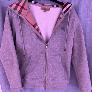 Grå zip-hoodie från Burberry - Säljer en grå hoodie från Burberry med dragkedja och klassiskt rutigt foder i huvan. Hoodien har två fickor fram, ribbade muddar och snören i huvan. Materialet är mjukt och bekvämt, perfekt för en avslappnad stil. Ikoniska Burberry-detaljer gör den extra snygg. Hör av er om ni undrar något🤝😄