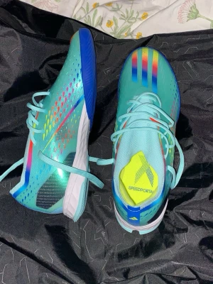 Adidas Speedportal turkosa fotbollsskor - Snygga Adidas Speedportal fotbollsskor i turkos med regnbågsfärgade detaljer och blå sula. Skorna har snörning och är tillverkade i syntetmaterial för bästa prestanda på planen. Perfekta för dig som vill sticka ut med både stil och fart. Storlek 39,5 aldrig använda