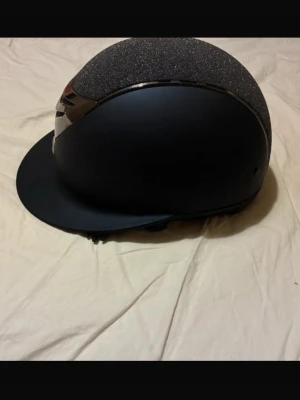 Svart ridhjälm från OneK - Snygg svart ridhjälm från OneK med glittrig ovandel och detaljer i silver. Hjälmen har en matt finish och justerbart hakband i vadderat material för extra komfort. Perfekt för ridning och tävling storlek 53-54 om jag minns rätt? Går att byta ilägg i den .
