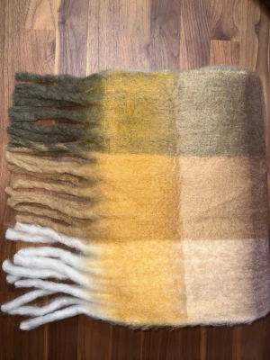 Rutig halsduk i brunt, beige och vitt - Mjuk och fluffig halsduk med rutigt mönster i brunt, beige och vitt. Halsduken har långa fransar i båda ändar och är perfekt för att hålla värmen under kalla dagar. Materialet känns ulligt och ger en mysig känsla. Halsduken liknar ”Acne Studios halsdukarna och är oanvänd (ny).