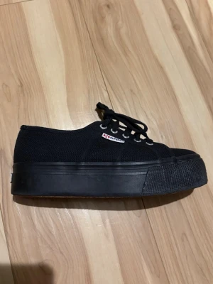 Svarta Superga sneakers med platåsula - Klassiska svarta Superga sneakers med platåsula. Skorna har snörning och är tillverkade i canvas med gummisula. 