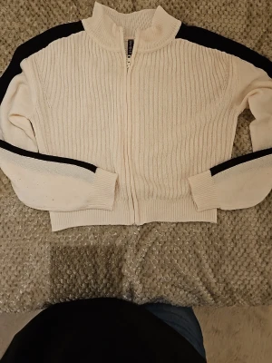 Vit stickad ziptröja från H&M Divided - Vit ribbstickad ziptröja från H&M Divided med svarta ränder längs ärmarna. Tröjan har hög krage, dragkedja framtill och långa ärmar. Perfekt för en avslappnad och sportig look.
