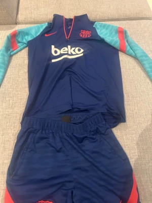 Barça träningsset Nike blå/röd - Snyggt träningsset från FC Barcelona och Nike. Tröjan är blå med ljusblå ärmar och röda detaljer, halv dragkedja och klubbmärke på bröstet. Matchande blå shorts med röda inslag och elastisk midja. Perfekt för fotbollsträning eller gymmet. Anledning till att jag inte använder denna mer är för att den är för liten.