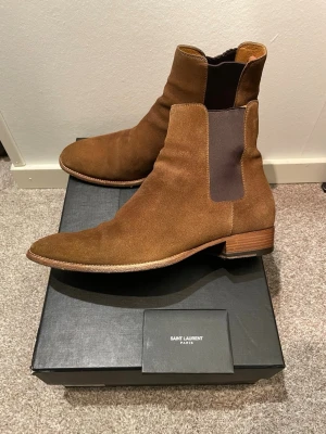 Saint Laurent Wyatt Hedi Slimane boots - Säljer dessa Saint Laurent Wyatt Chelsea Boots, designade av den berömde Hedi Slimane. Köpta från AFound och orderbekräftelse finns! Säljer pga jag tyvärr inte gillade färgen. Klackens höjd 30mm samt innersulans mått är 29cm. Stl 40 men sitter stort i storleken. Tillverkade i högkvalitativt kalv läder med en unik design, dessa skor är till för dig som vill ha det allra bästa. Hör av dig så fort du undrar något!! Nypris 12 000kr