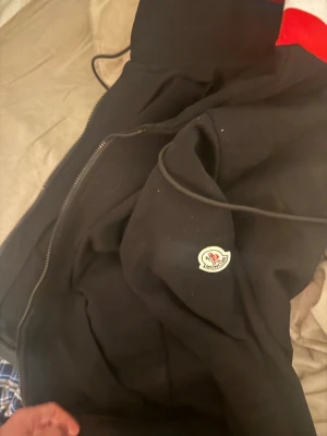 Svart Moncler hoodie med dragkedja - Svart hoodie från Moncler med dragkedja framtill och klassisk logotyp på bröstet. Huvan har snygga ränder i rött, vitt och blått. Perfekt för dig som vill ha en stilren och exklusiv look.