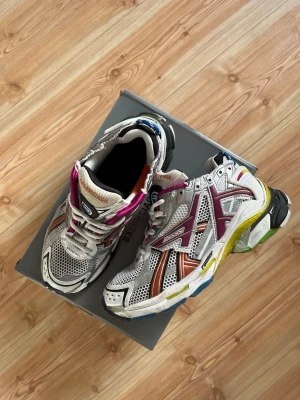 BaIenciaga Runner sneakers multicolor - Säljer ett par BaIenciaga Runner sneakers i mesh och syntetmaterial. Skorna är silvriga med detaljer i lila, orange, blått, svart och gult. De har en chunky sula och sportig design med flera färginslag och logga på sidan. Perfekta för dig som gillar statement-sneakers.