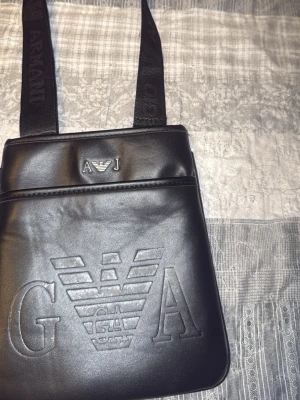 Svart axelväska från Armani Jeans - Snygg svart axelväska från Armani Jeans i skinnimitation med präglad logga framtill och justerbar axelrem med textdetaljer. Perfekt för att bära det viktigaste och ger en clean och stilren look.