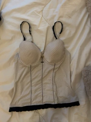 Vit korsett topp med svarta prickar och spets - Snygg vit korsett/topp med svarta prickar, axelband och svart spetskant nedtill. Kuporna är vadderade och tyvärr lite små för mig, därför jag säljer🥲går att klä upp och ner den, skitsnygg till både fest och vardags!! står ingen storlek men skulle säga s