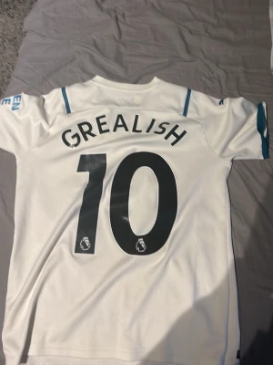 Manchester City Grealish tröja Puma - Säljer en vit Manchester City matchtröja från Puma med Grealish och nummer 10 på ryggen. Tröjan har korta ärmar, v-ringning och klubbmärke samt Etihad Airways-tryck på bröstet. Perfekt för fotbollsfans som vill stötta sitt lag.