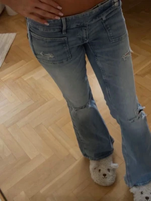 Blå bootcut jeans low Rise! - Säljer ett par ljusblå bootcut jeans med slitningar och hål vid knäna. Jeansen har låg midja, klassiska fem fickor och en avslappnad passform. Perfekta för en trendig och avslappnad look.