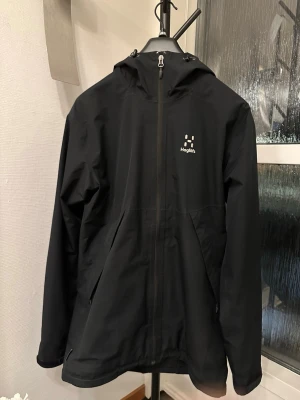 Svart vindjacka från Haglöfs - Snygg svart vindjacka från Haglöfs med justerbar huva. Jackan bär storleken S men eftersom den är en lite längre modell av haglöfs så passar den också M. Personligen bär jag S/M och upplever att den sitter bra. Jackan har hel dragkedja framtill,även justerbar vid armen. meshfoder på insidan för extra ventilation. Perfekt för blåsiga dagar och outdoor-äventyr. Skick 7/10, finns små repor vid armen men syns knappt annars är den 9/10. Nypris : 1900kr (pris kan diskuteras vid snabb affär).