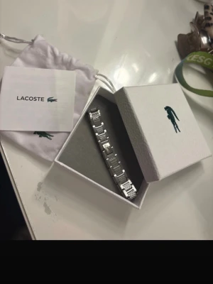 Silverfärgad klocka från Lacoste - Snygg och stilren armband från Lacoste i silverfärgat metallarmband. Kommer i originalförpackning med tillhörande påse och certifikat. Perfekt accessoar för dig som gillar klassisk och modern design med Lacostes ikoniska krokodillogga.