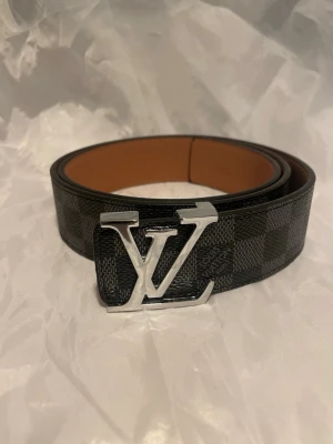 Svart grått bälte med silver spänne. 110cm - Snyggt skärp med rutigt mönster i svart och grått. Skärpet har ett stort silverfärgat spänne. 110cm pris går att diskutera 