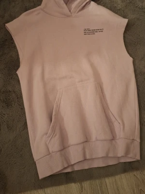 Ljusrosa ärmlös hoodie med tryck - Säljer en ljusrosa ärmlös hoodie med huva och magficka. På bröstet finns ett svart tryck med texten 'new york / THE CITY WHERE DREAMS BECOME REALITY.' Hoodien är i mjukt sweatshirtmaterial och har ribbad nederkant. Perfekt för en chill och avslappnad stil.