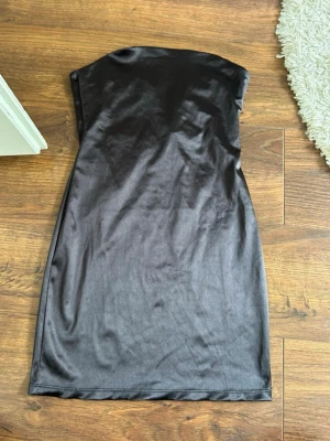 Svart axelbandslös satinklänning - Snygg svart axelbandslös klänning i satinliknande material. Kort modell med figurnära passform och stilren design, perfekt för dig som gillar en enkel men elegant look.