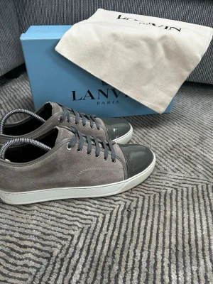 Lanvin skor gråa  - Snygga sneakers från Lanvin i grå mocka med mörkgrå lackad tå och matchande grå skosnören. Skorna har vit sula och klassisk låg profil. Perfekta för dig som vill ha en stilren och exklusiv look.