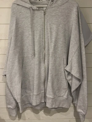 Ljusgrå zip-hoodie med huva - Snygg ljusgrå hoodie med dragkedja framtill och två stora fickor. Modellen har huva med dragsko och ribbade muddar vid ärmslut och nederkant. Perfekt för en avslappnad och chill stil. Materialet är mjukt och skönt, troligtvis bomullsblandning.